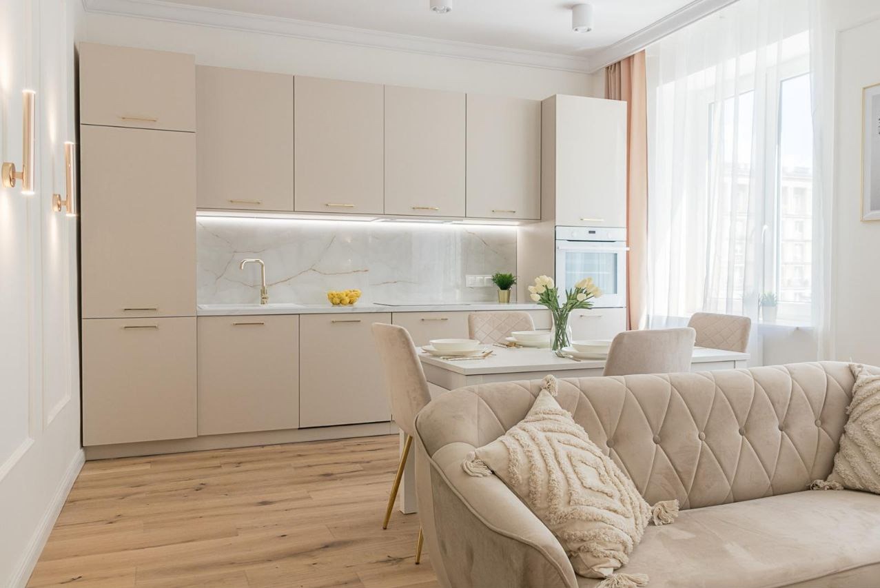 Jasna kuchnia z beżowymi szafkami, marmurowym blatem i złotymi akcentami, widok z salonu z beżową sofą i poduszkami z frędzlami.
