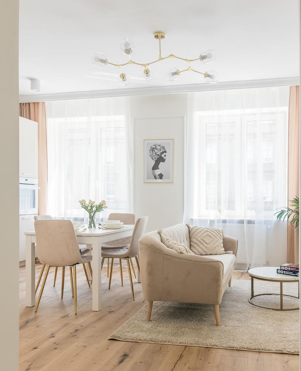 Jasne wnętrze salonu z jadalnią: drewniana podłoga, biały stół z beżowymi krzesłami na złotych nogach, beżowa sofa, okrągły stolik kawowy, białe zasłony i lampa sufitowa w stylu retro.