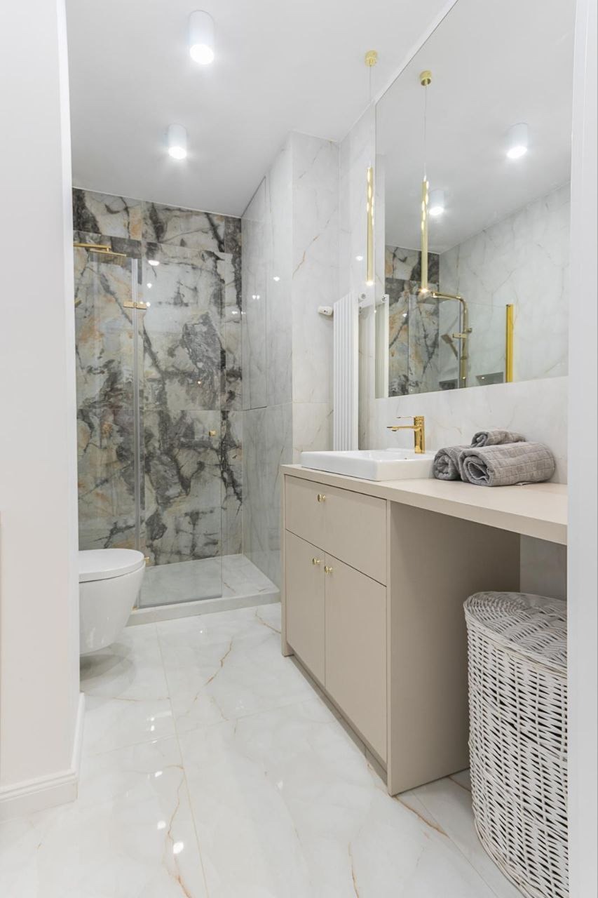 Jasna łazienka z marmurowymi płytkami w odcieniach szarości i złota, kabina prysznicowa ze złotymi okuciami, beżowa szafka z umywalką i złotą armaturą, dekoracyjne lampy wiszące, biała podłoga...