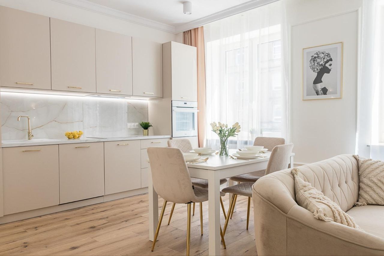 Jasna kuchnia z jadalnią w Warszawie. Kremowe szafki, marmurowy backsplash, stół z białym blatem i welurowe krzesła na złotych nogach. Na stole zastawa i bukiet białych tulipanów.