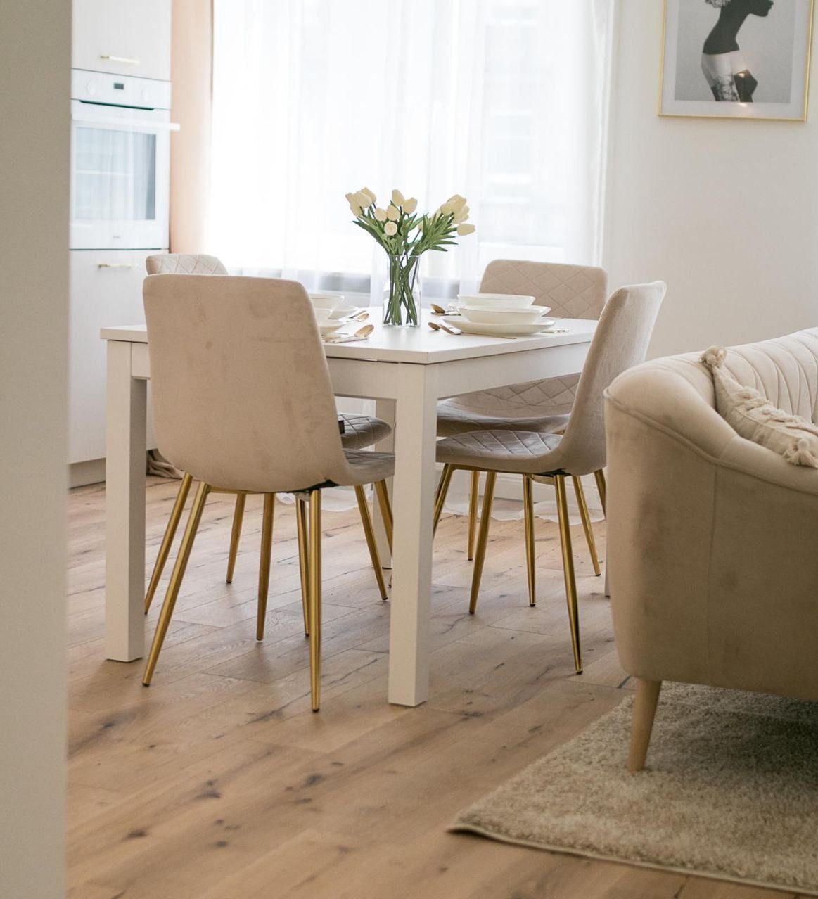Jasna jadalnia z białym stołem, beżowymi krzesłami na złotych nogach, wazonem z tulipanami i fragmentem beżowej sofy, na podłodze parkiet.