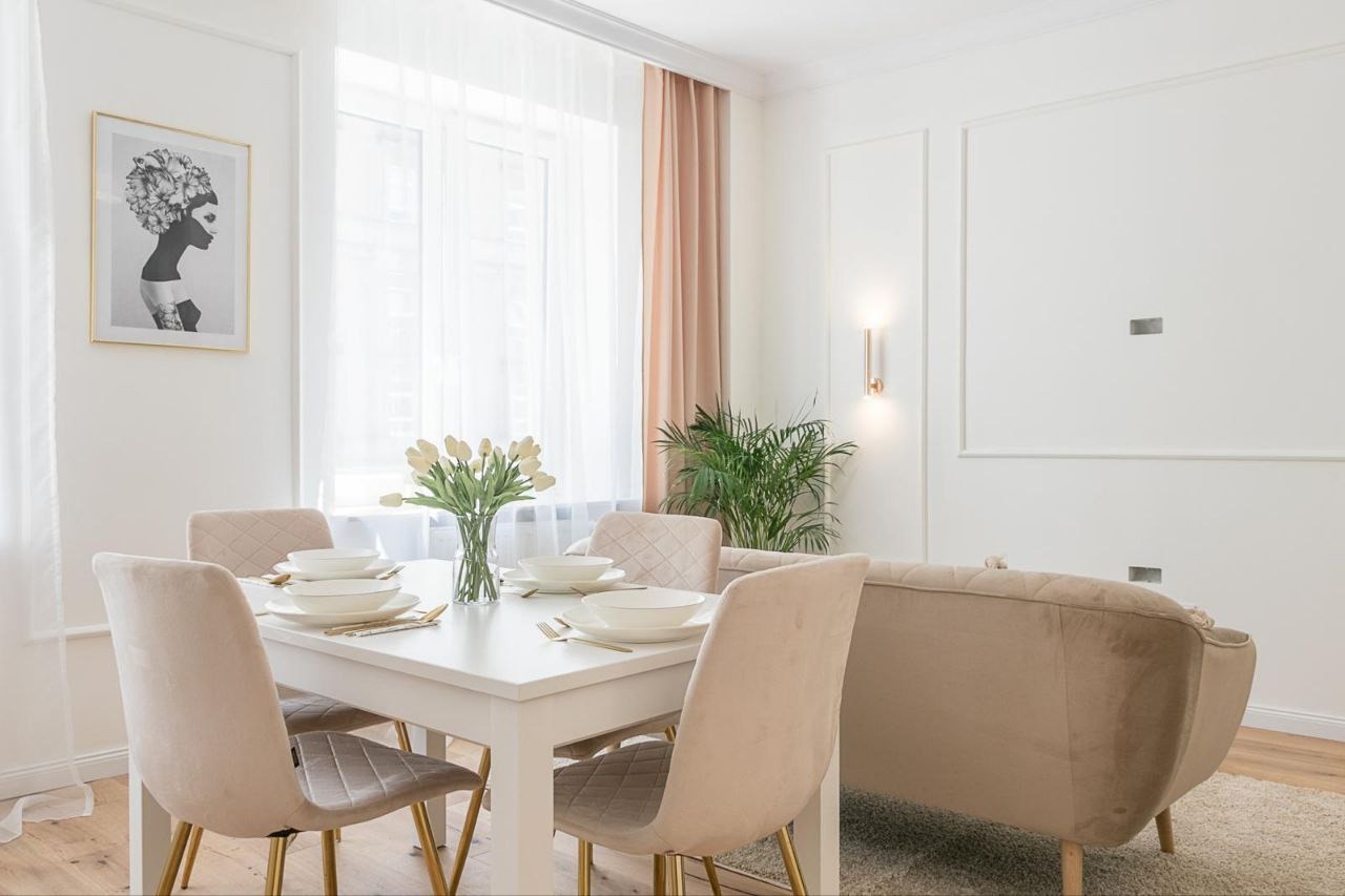 Jasne wnętrze jadalni z białym stołem nakrytym dla czterech osób, beżowe krzesła z pikowaniem, sofa w odcieniach beżu, na ścianie grafika w złotej ramie i minimalistyczne kinkiety.