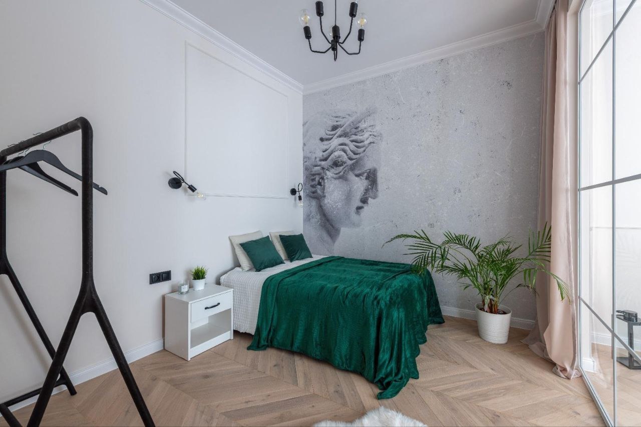 Jasna sypialnia z parkietem w jodełkę, ścianą z motywem rzeźby, łóżkiem przykrytym szmaragdowym pledem i czarnym wieszakiem na ubrania.