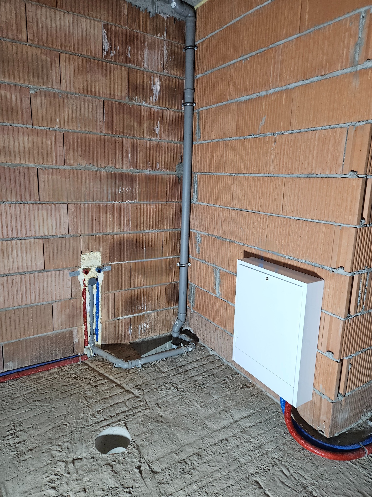 Wnętrze w trakcie prac hydraulicznych: widoczne surowe ściany z cegły, pionowa rura kanalizacyjna z uchwytami, wystające rury ciepłej i zimnej wody zabezpieczone pianką oraz biała skrzynka...