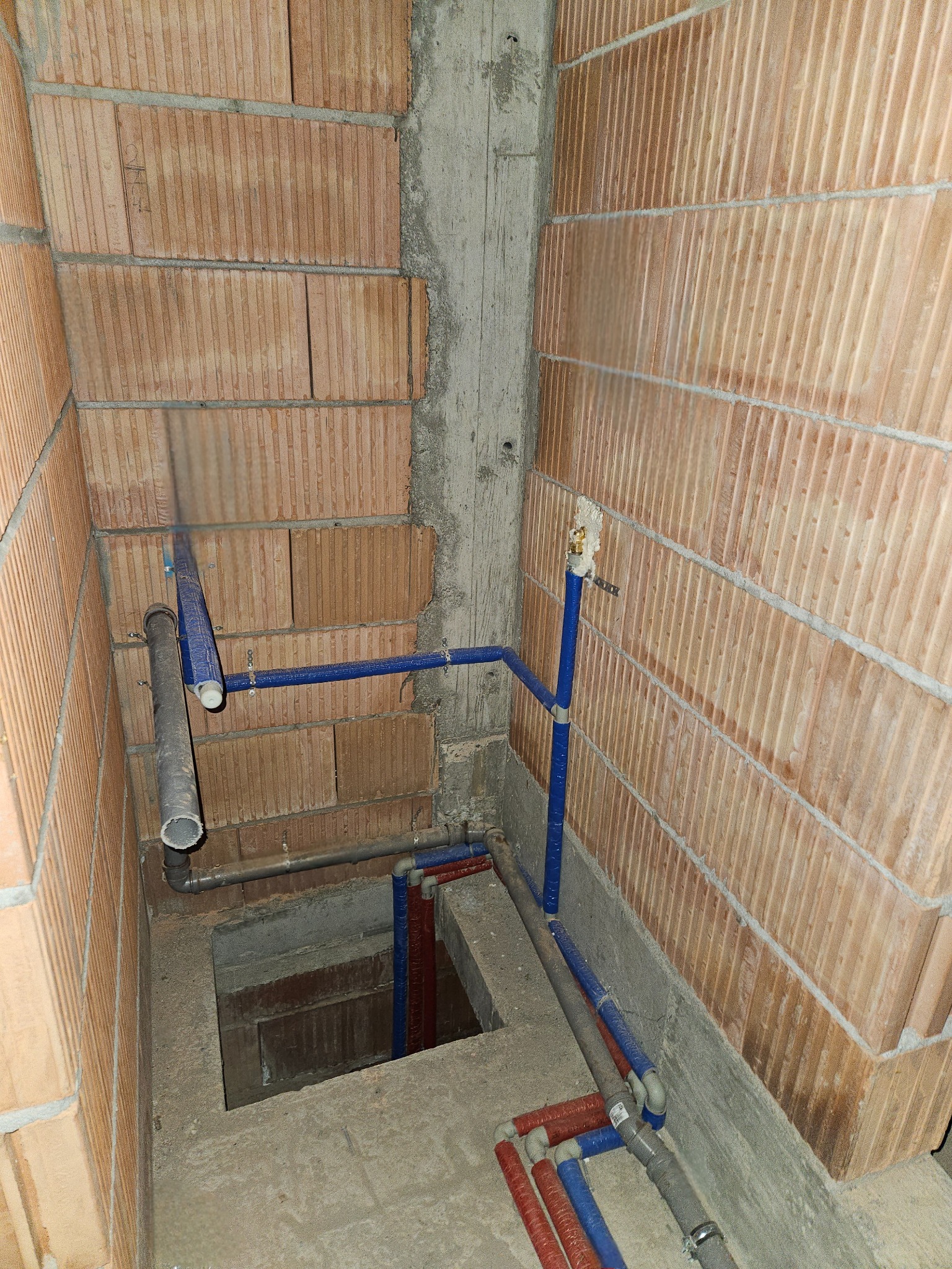 Surowe wnętrze w trakcie budowy z widocznymi instalacjami wodno-kanalizacyjnymi: rury niebieskie i szare prowadzone wzdłuż ceglanej ściany i w dół do otworu w posadzce.