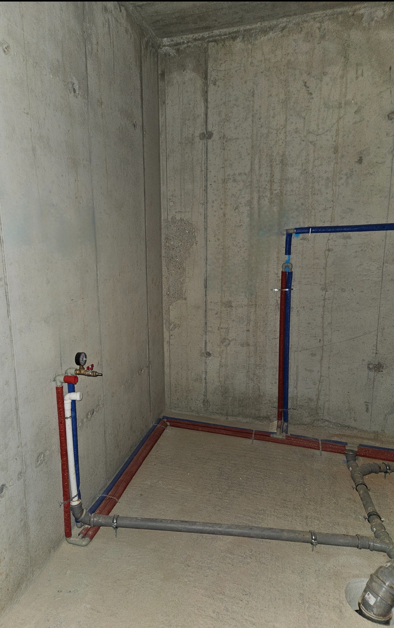Surowy betonowy kąt piwnicy z nową instalacją hydrauliczną: rury PEX w kolorze czerwonym i niebieskim ułożone wzdłuż ścian, szare rury kanalizacyjne z metalowymi obejmami na podłodze, manometr przy...