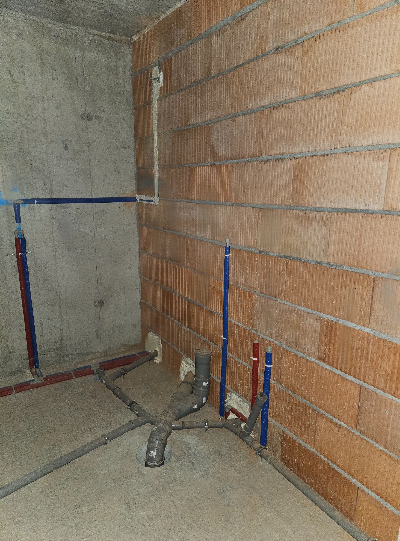 Surowy stan: Instalacja kanalizacyjna z szarych rur PCV w narożniku pomieszczenia z ceglanymi i betonowymi ścianami, widoczne niebieskie i czerwone rury doprowadzające wodę.