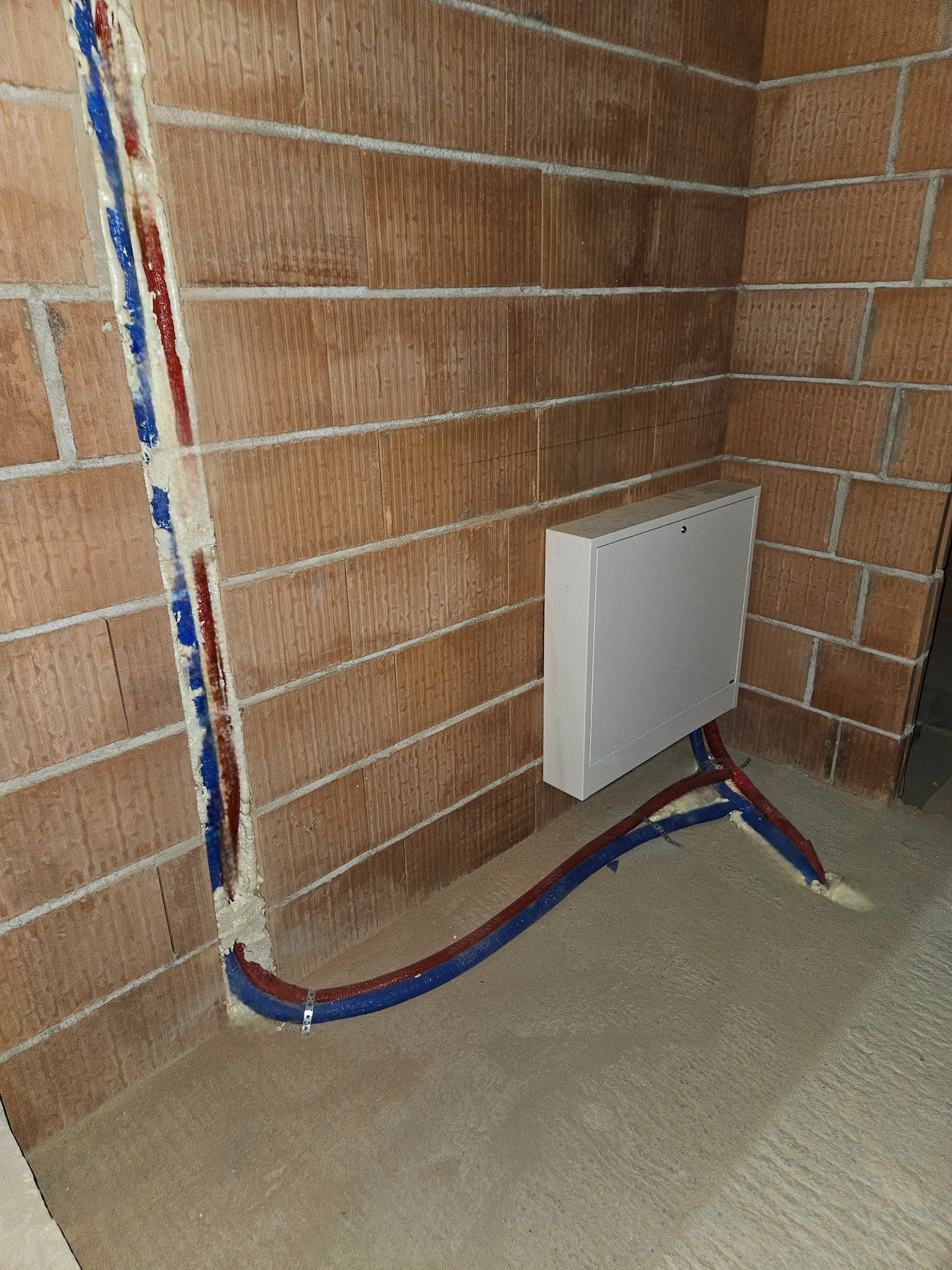Wewnętrzna instalacja wodna z rurami w kolorach niebieskim i czerwonym wychodzącymi ze ściany z cegły, zabezpieczone pianką montażową i przymocowane do podłoża, z widoczną białą skrzynką rozdzielczą.