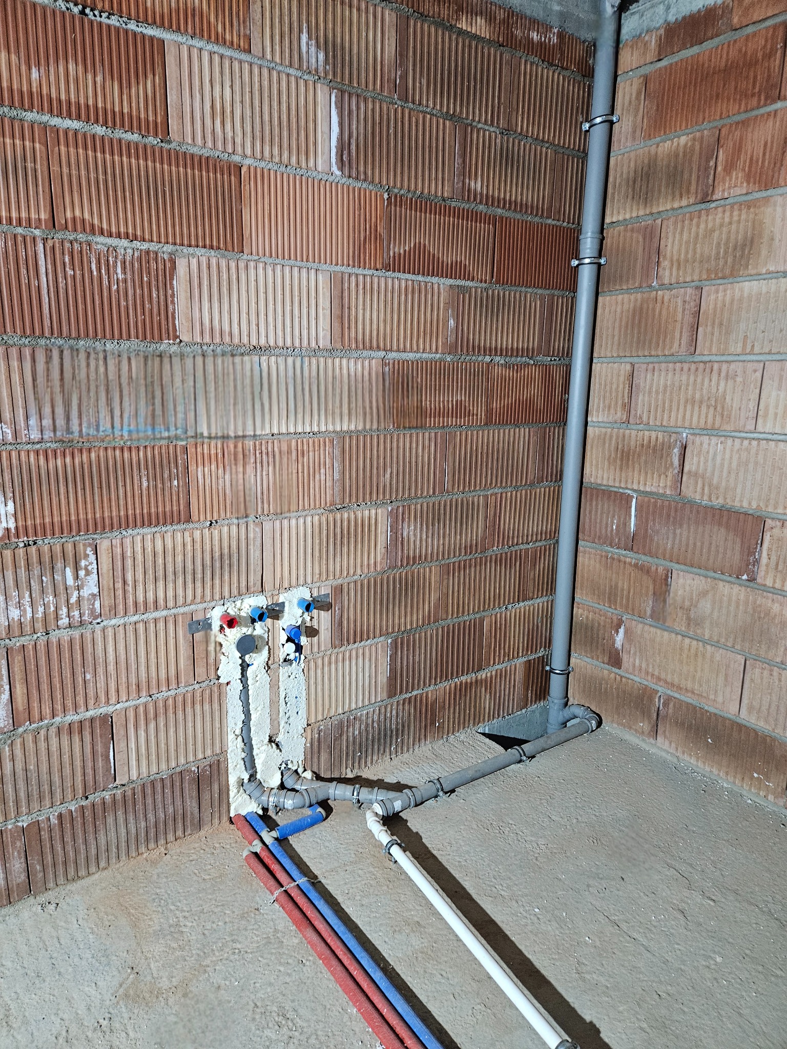 Surowe wnętrze z widocznymi instalacjami hydraulicznymi: rury z tworzywa w kolorach czerwonym, niebieskim i białym, wychodzące ze ściany z cegły dziurawki, oraz szara rura odpływowa mocowana...