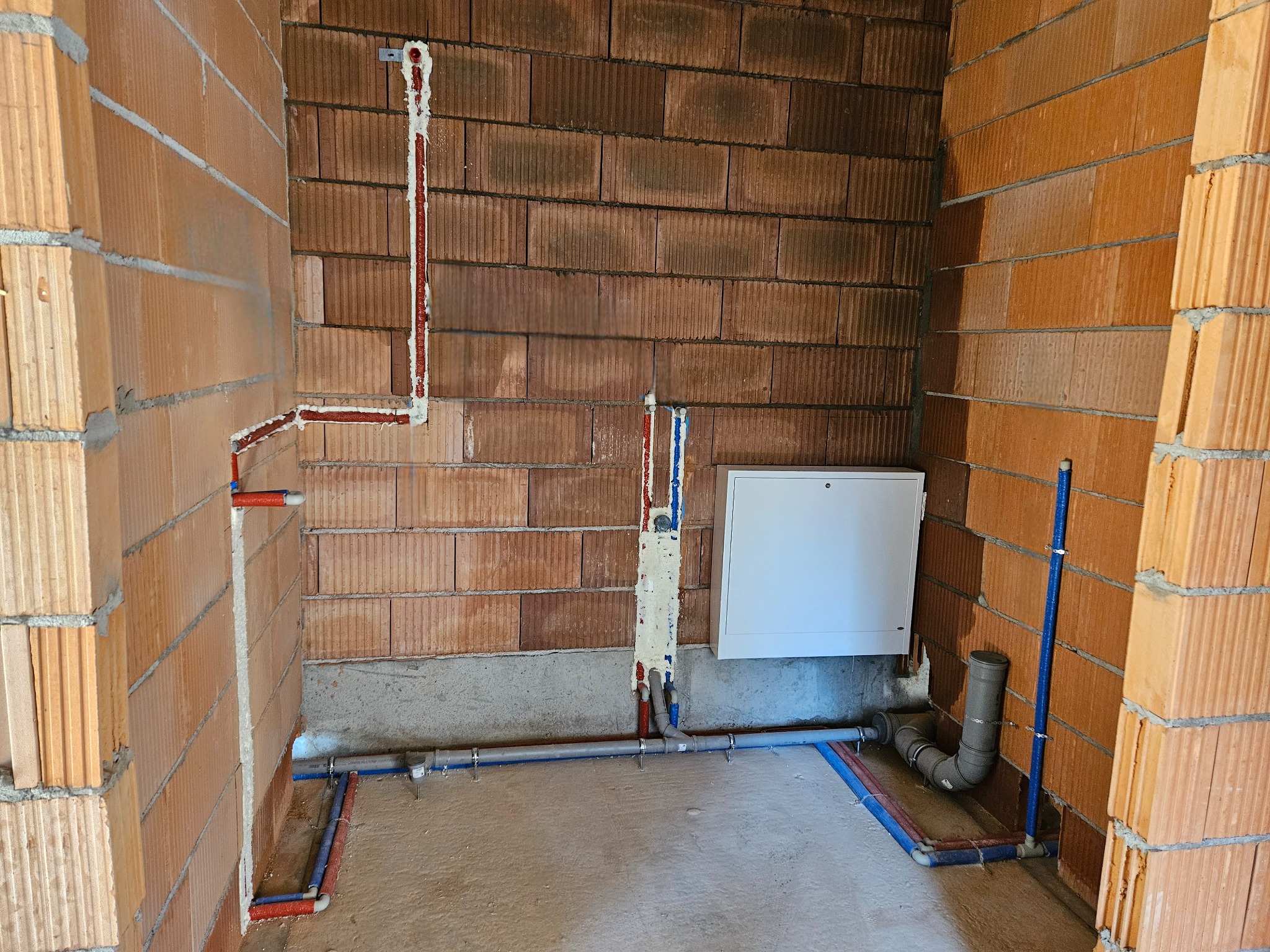 Surowe wnętrze z widocznymi instalacjami wodno-kanalizacyjnymi w trakcie montażu; rury z czerwonymi i niebieskimi paskami prowadzone po ścianach z cegły, częściowo osadzone w pianie montażowej...