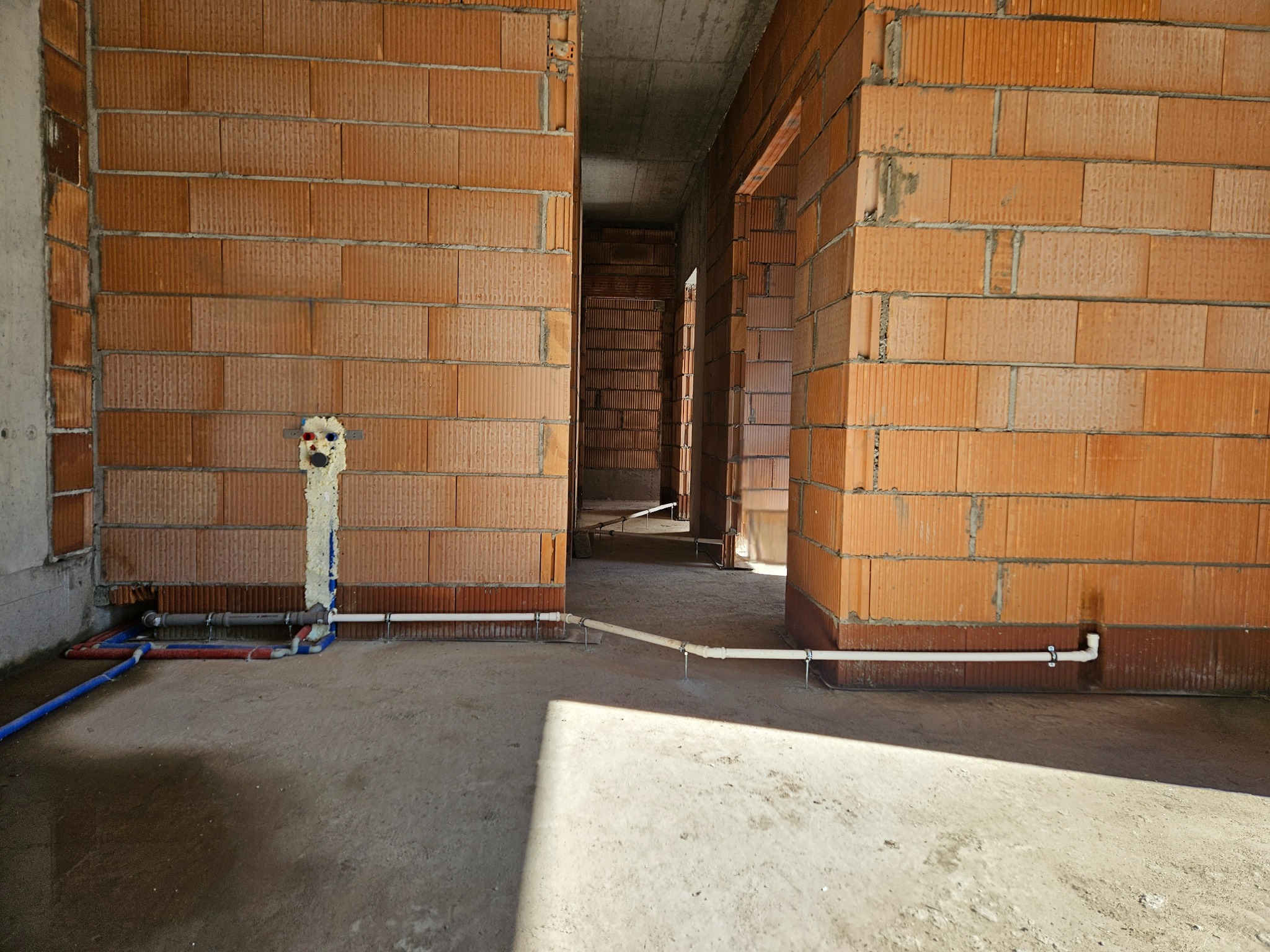 Surowy stan: wewnątrz budynku w trakcie budowy widoczne poprowadzone rury instalacji wodnej i kanalizacyjnej, zabezpieczone pianką montażową, na tle ścian z cegły dziurawki.