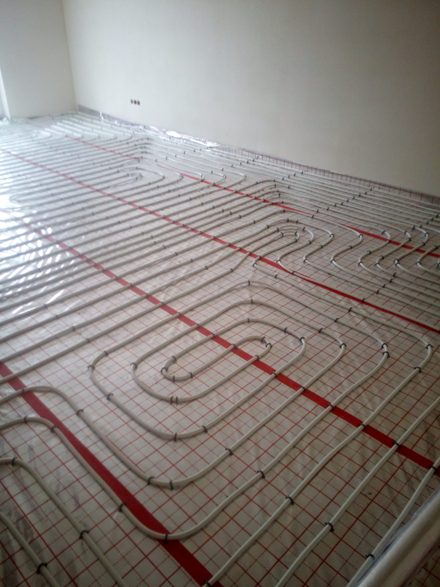 Białe rury ogrzewania podłogowego rozłożone na folii z czerwoną siatką w pustym pomieszczeniu, widoczne czarne zaciski mocujące rury.