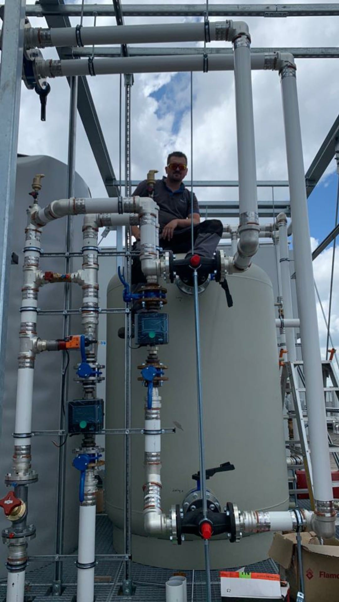 Instalator w okularach siedzi na zbiorniku wyrównawczym, kontrolując połączenia rur z zaworami i czujnikami w instalacji przemysłowej na zewnątrz budynku.
