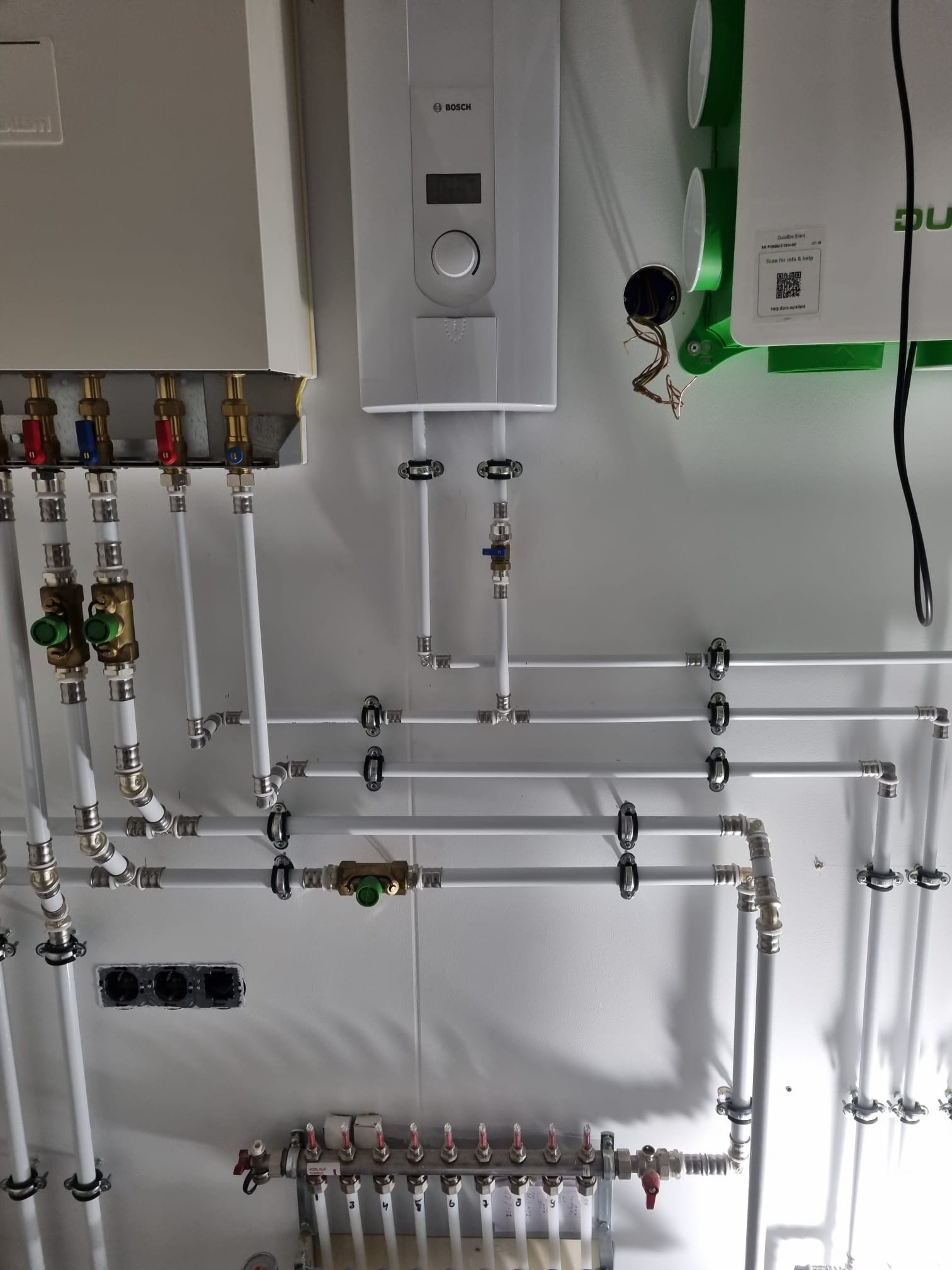 Rozbudowana instalacja hydrauliczna i grzewcza z białych rur, obejmująca rozdzielacz podłogowy, kotły Bosch, zawory i liczne połączenia, zamontowana na białej ścianie w kotłowni.