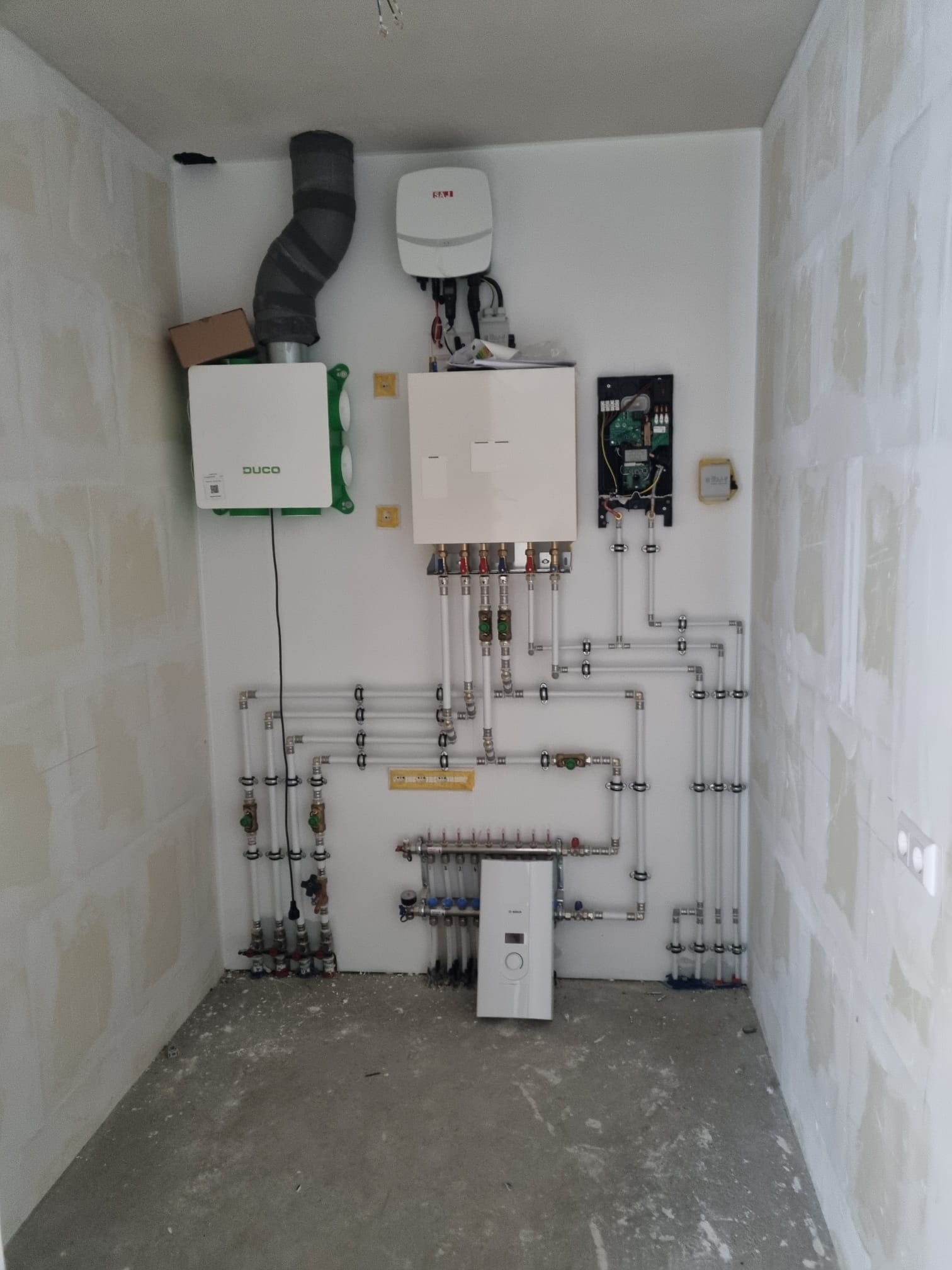 Wnętrze pomieszczenia z zamontowaną instalacją grzewczą: rekuperator, rozdzielacz, piec gazowy kondensacyjny i rozbudowany system rur z zaworami, ściany w trakcie wykańczania.