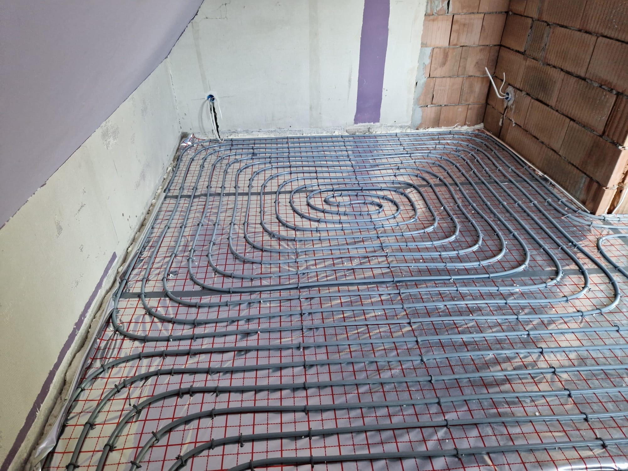 Szare rury ogrzewania podłogowego ułożone spiralnie na folii z czerwoną kratką w surowym pomieszczeniu z częściowo otynkowanymi ścianami i fragmentem ściany z cegły.