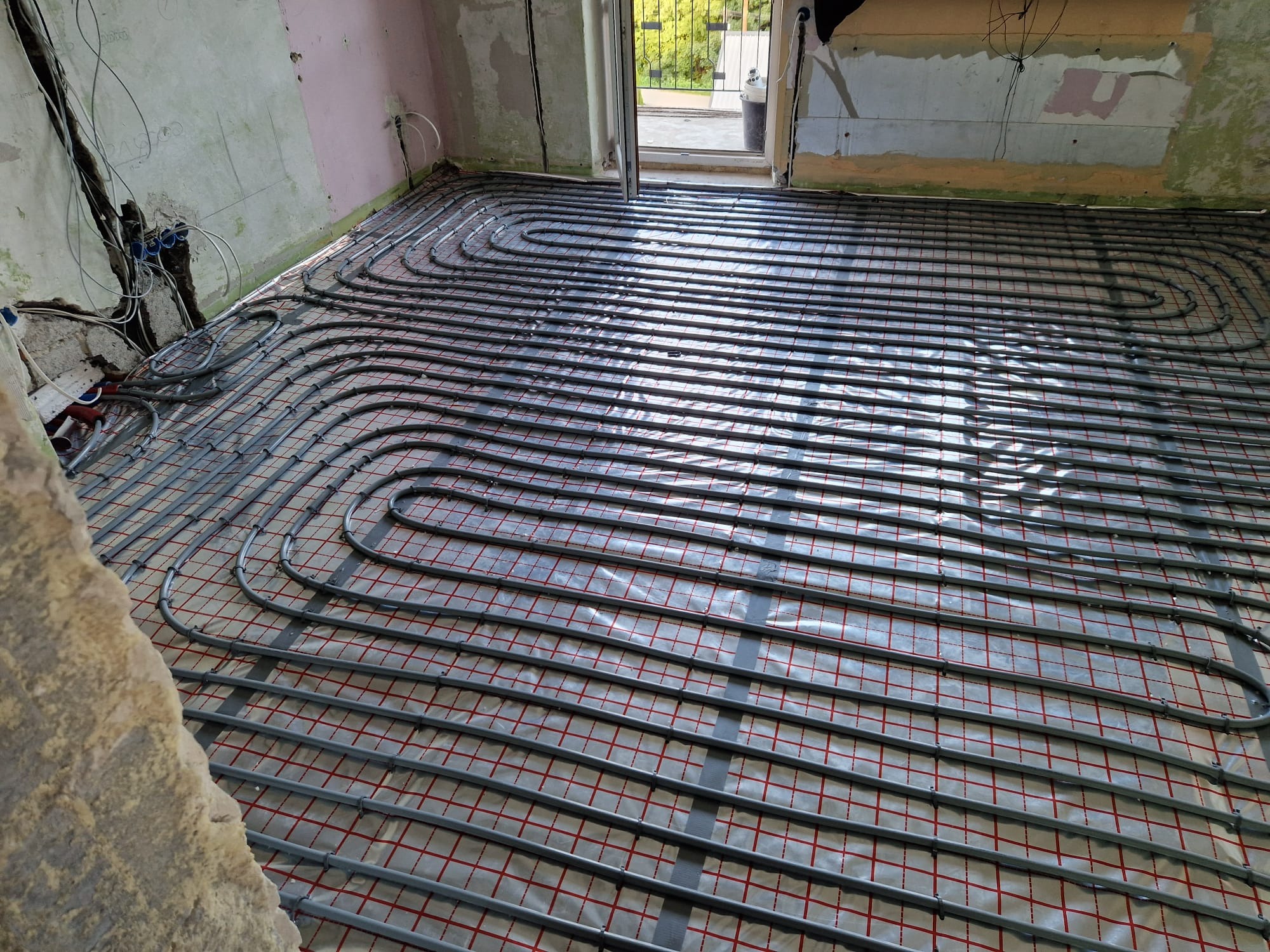 Instalacja ogrzewania podłogowego z szarych rur na folii z czerwoną kratką, widoczne nieotynkowane ściany i wystające kable.