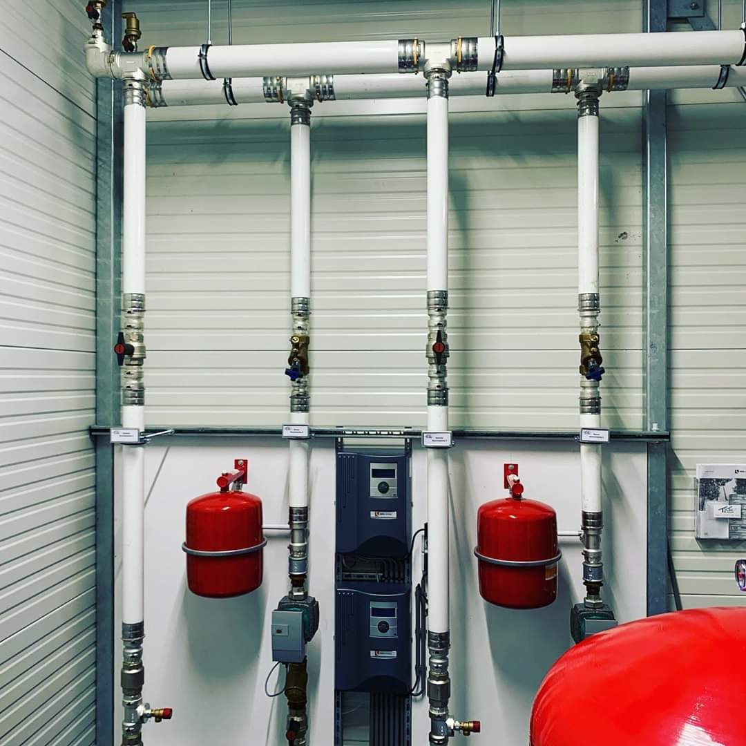 Rozbudowana instalacja centralnego ogrzewania z białymi rurami, czerwonymi zbiornikami wyrównawczymi i szarymi sterownikami, zamontowana na ścianie z blachy trapezowej.