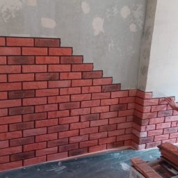 Lucerna Sp. z o.o. - Montaż dekoracyjnej cegły na ścianie wewnętrznej, widoczny fragment ściany surowej i częściowo wykończonej, pracownik dociska element do ściany, widoczne opakowania cegieł i czerwony mop.