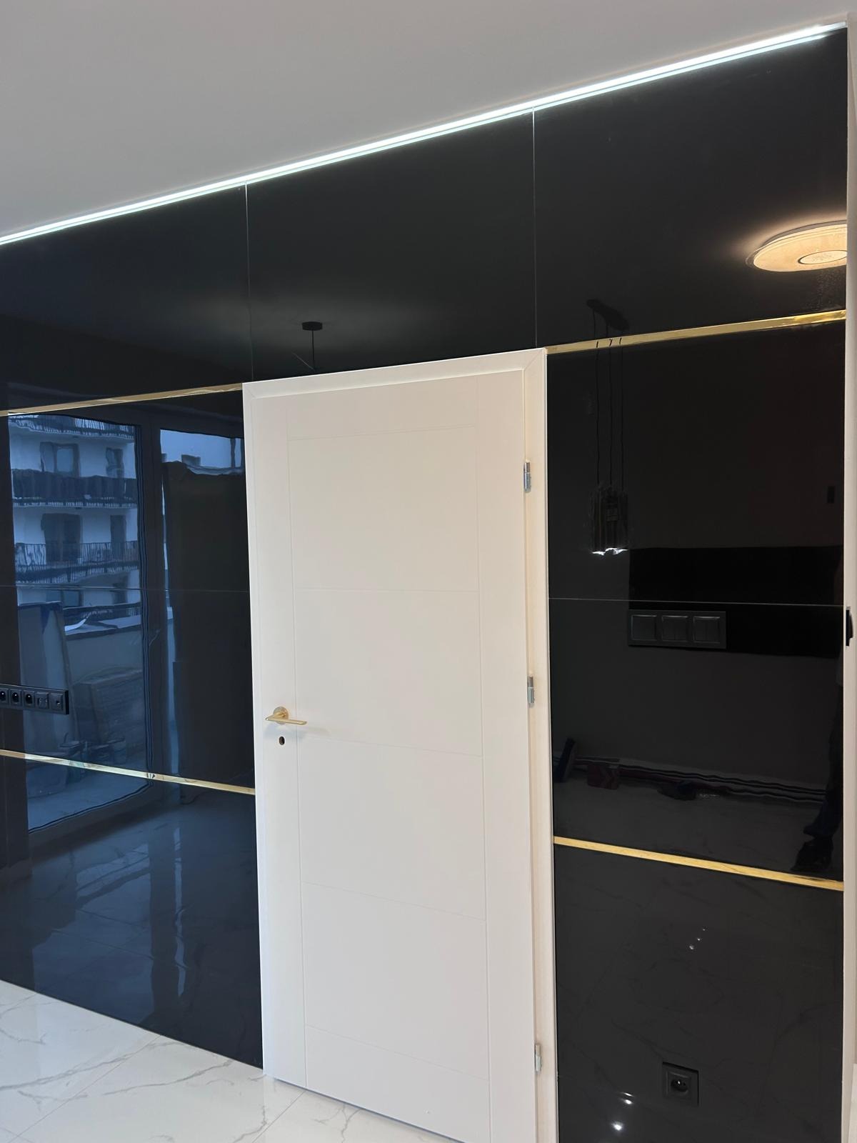 Nowoczesne wnętrze z białą, minimalistyczną ościeżnicą i drzwiami na tle błyszczącej czarnej ściany z dekoracyjnymi złotymi listwami, oświetleniem LED przy suficie i marmurową podłogą.