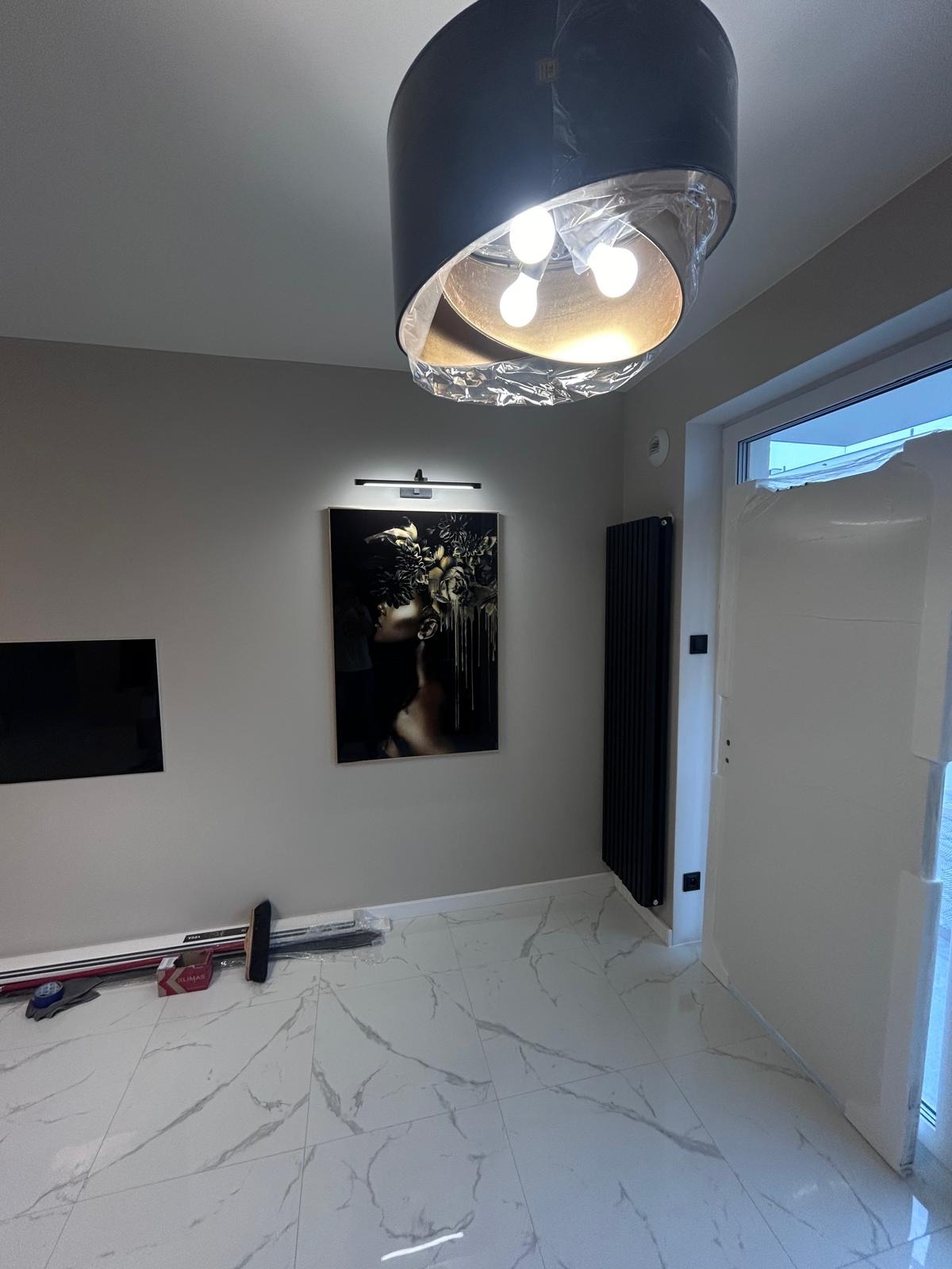Nowoczesne wnętrze z marmurową podłogą, obrazem z podświetleniem i lampą wiszącą w folii ochronnej, widoczne narzędzia przy ścianie.