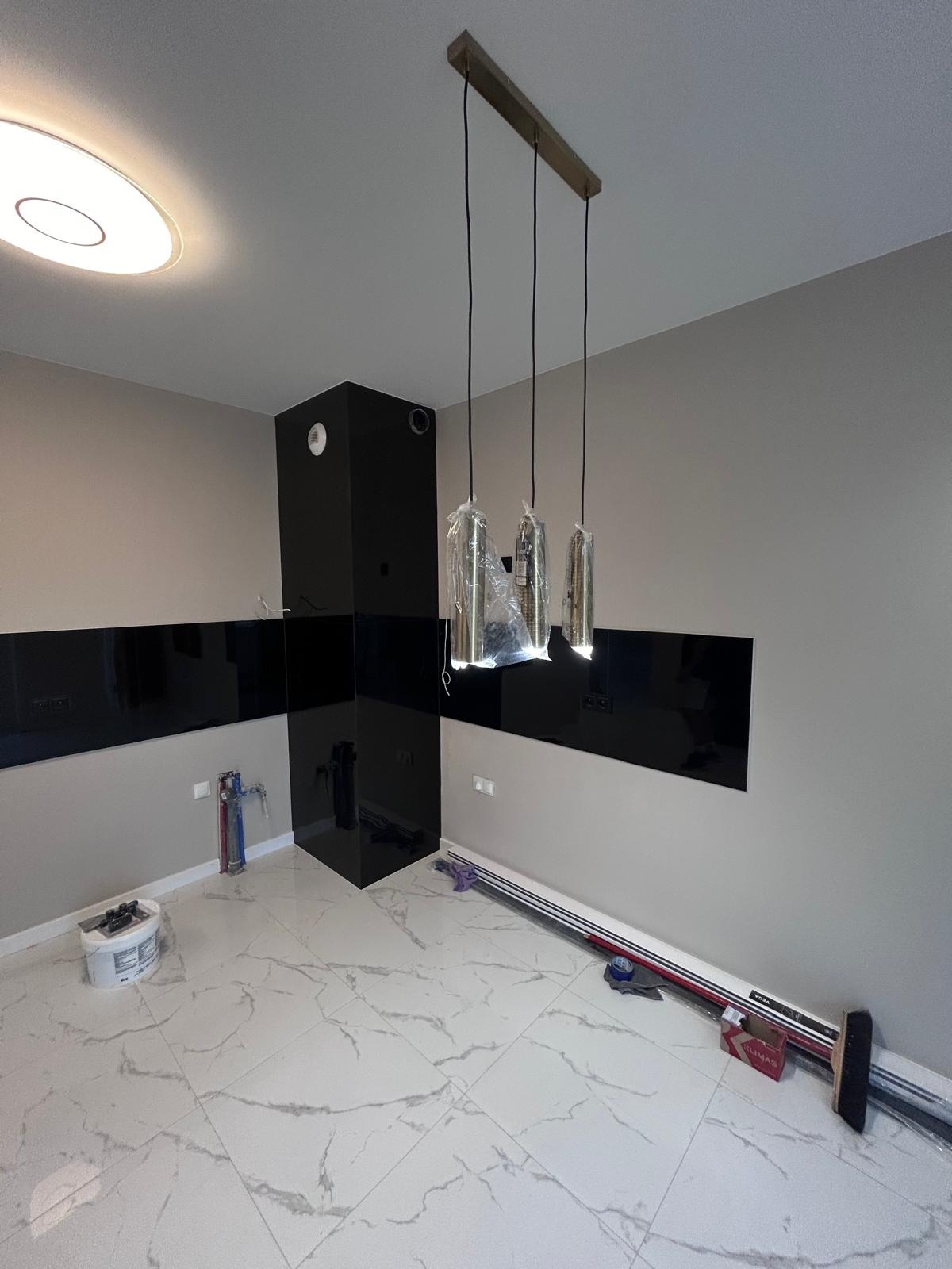Nowoczesna kuchnia w trakcie wykańczania: czarne panele szklane na ścianach, marmurowe płytki podłogowe, wiszące lampy w folii ochronnej, widoczne narzędzia i materiały budowlane.