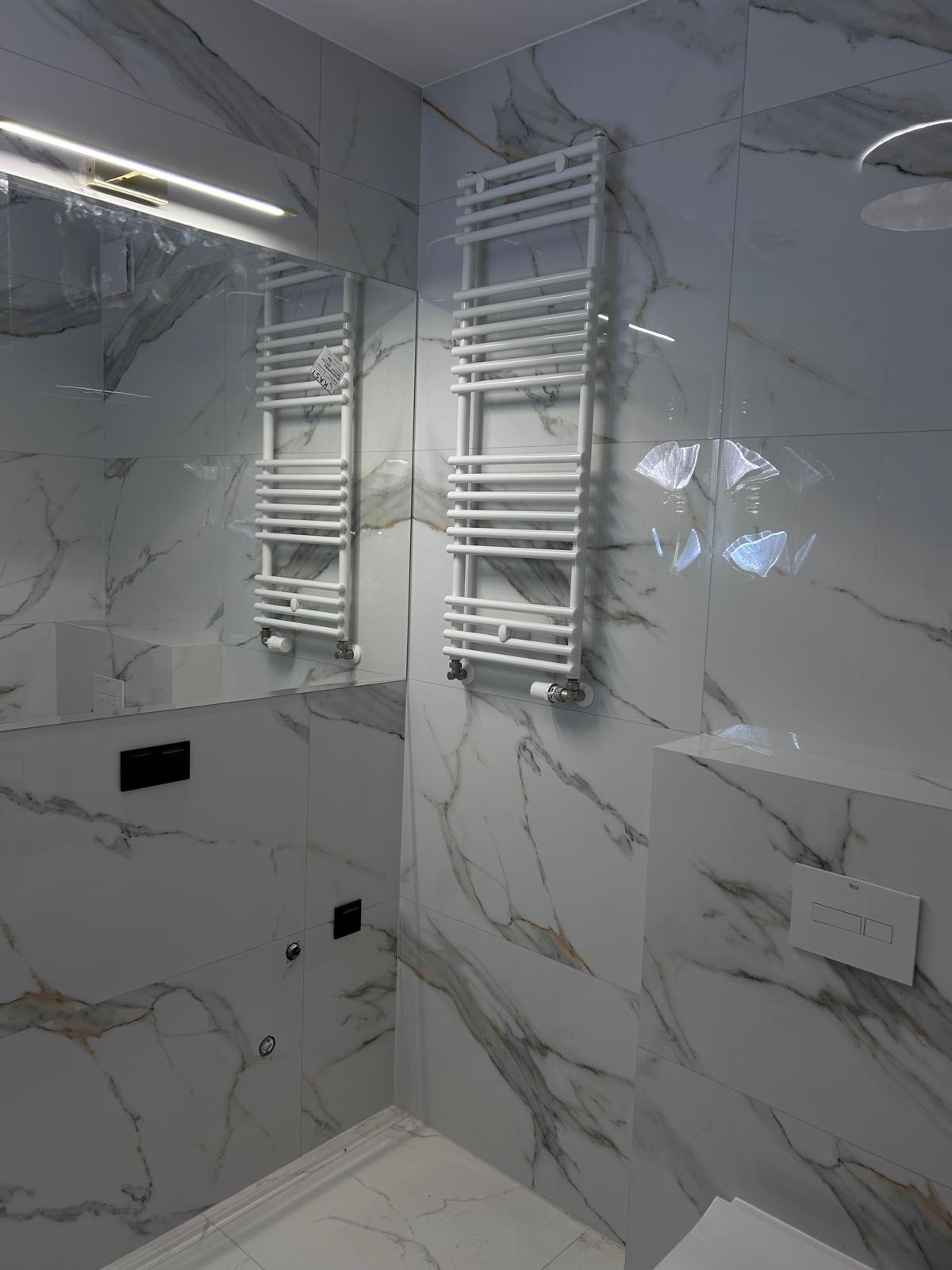 Biała łazienka z marmurowymi płytkami, grzejnik drabinkowy i podświetlane lustro.
