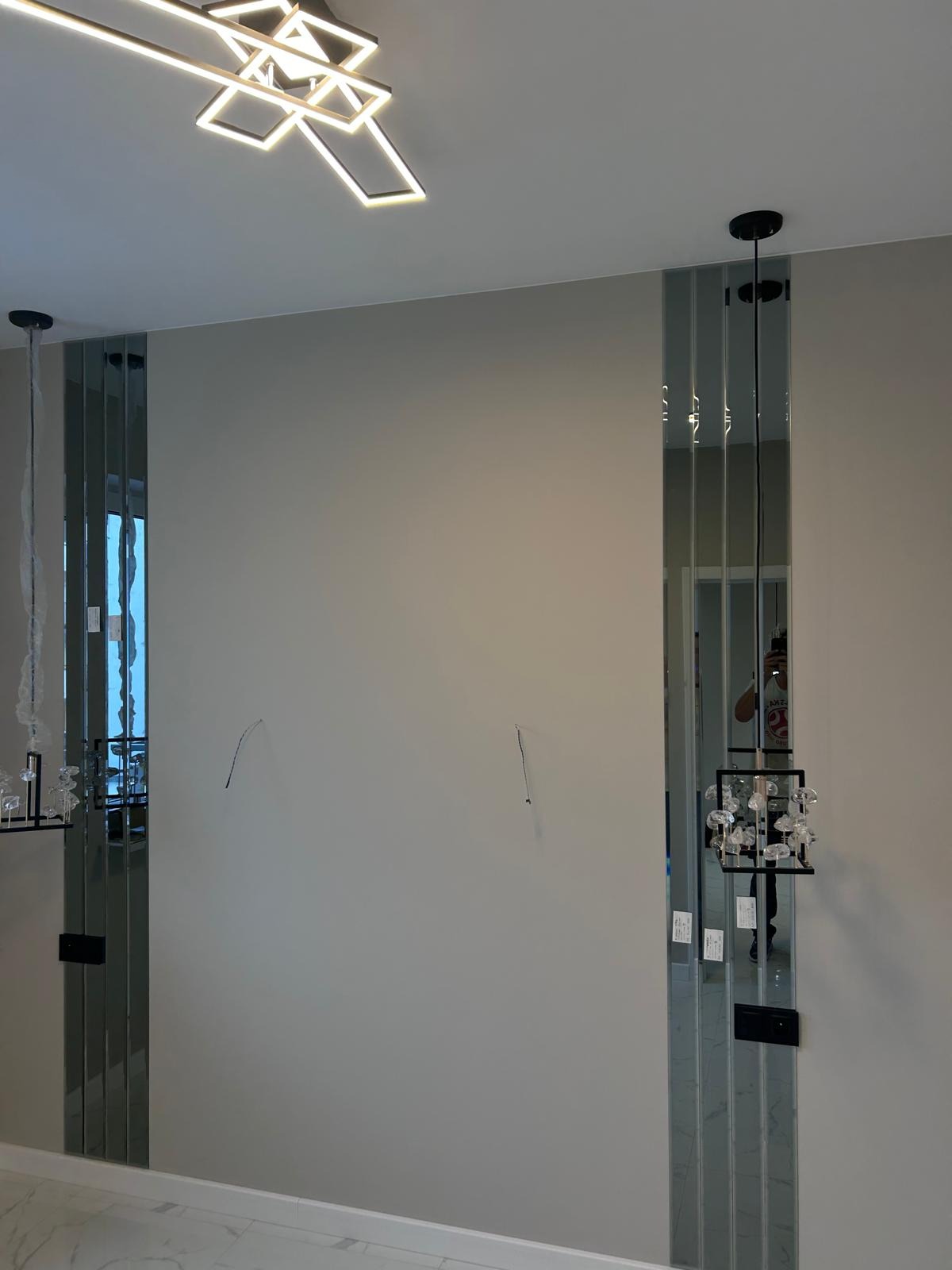 Ściana z zamontowanymi pionowymi pasami lustrzanymi po bokach, widoczne otwory po kablach i nowoczesna lampa sufitowa.