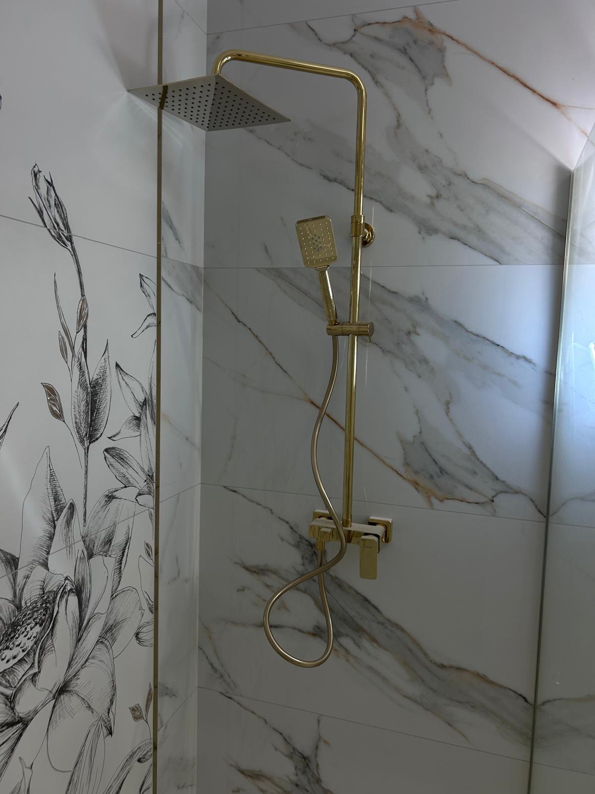 Złoty zestaw prysznicowy: deszczownica i słuchawka, zamontowany na ścianie z płytek imitujących marmur, obok ściany z motywem kwiatowym.