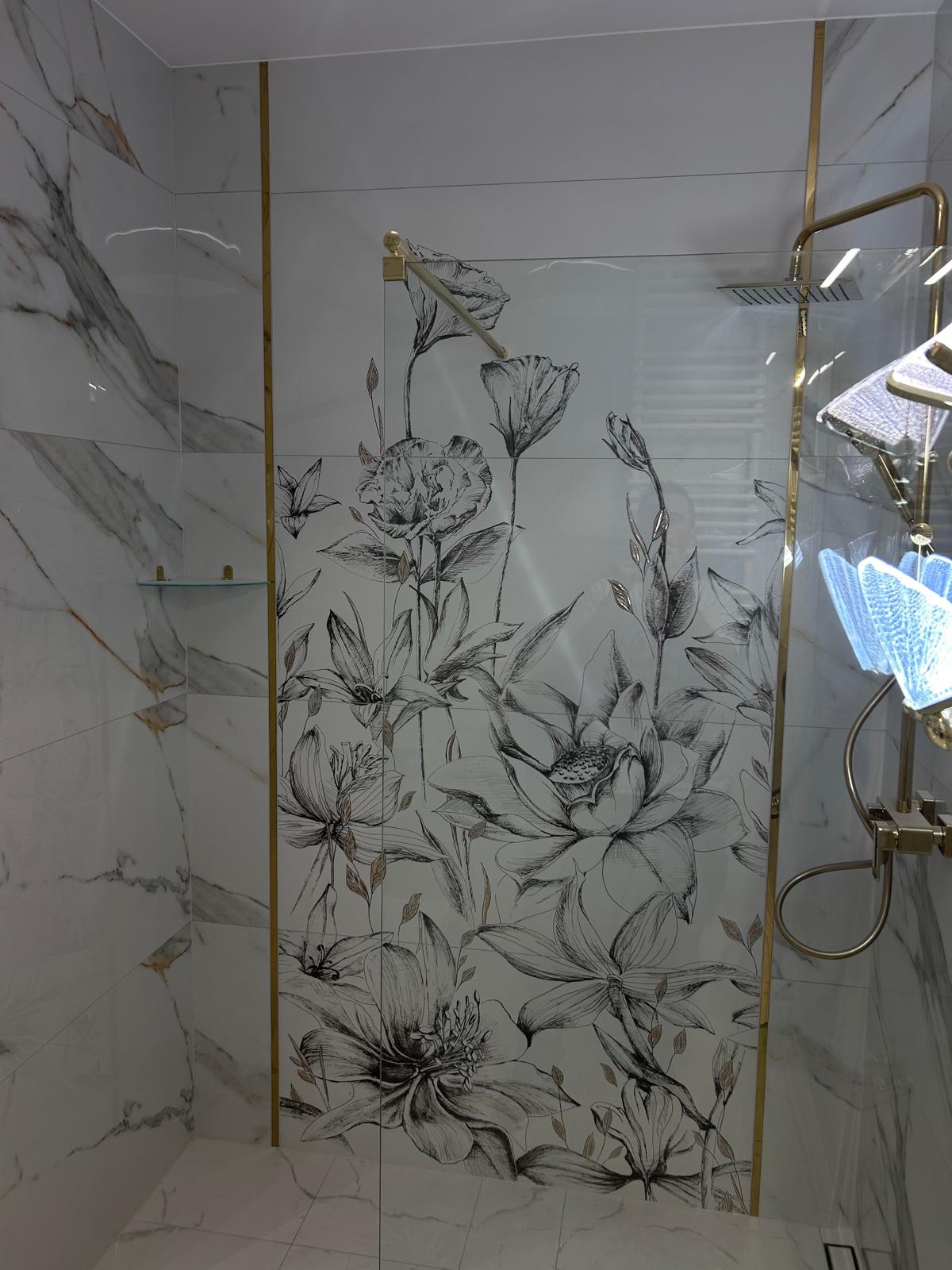 Wykończona łazienka z kabiną prysznicową, białe marmurowe płytki z motywem kwiatowym, złote wykończenia i deszczownica.