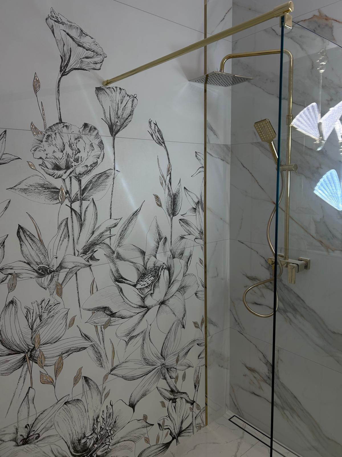Nowoczesna kabina prysznicowa z deszczownicą i baterią w kolorze złotym, na ścianach białe płytki z motywem kwiatowym w stylu szkicu.