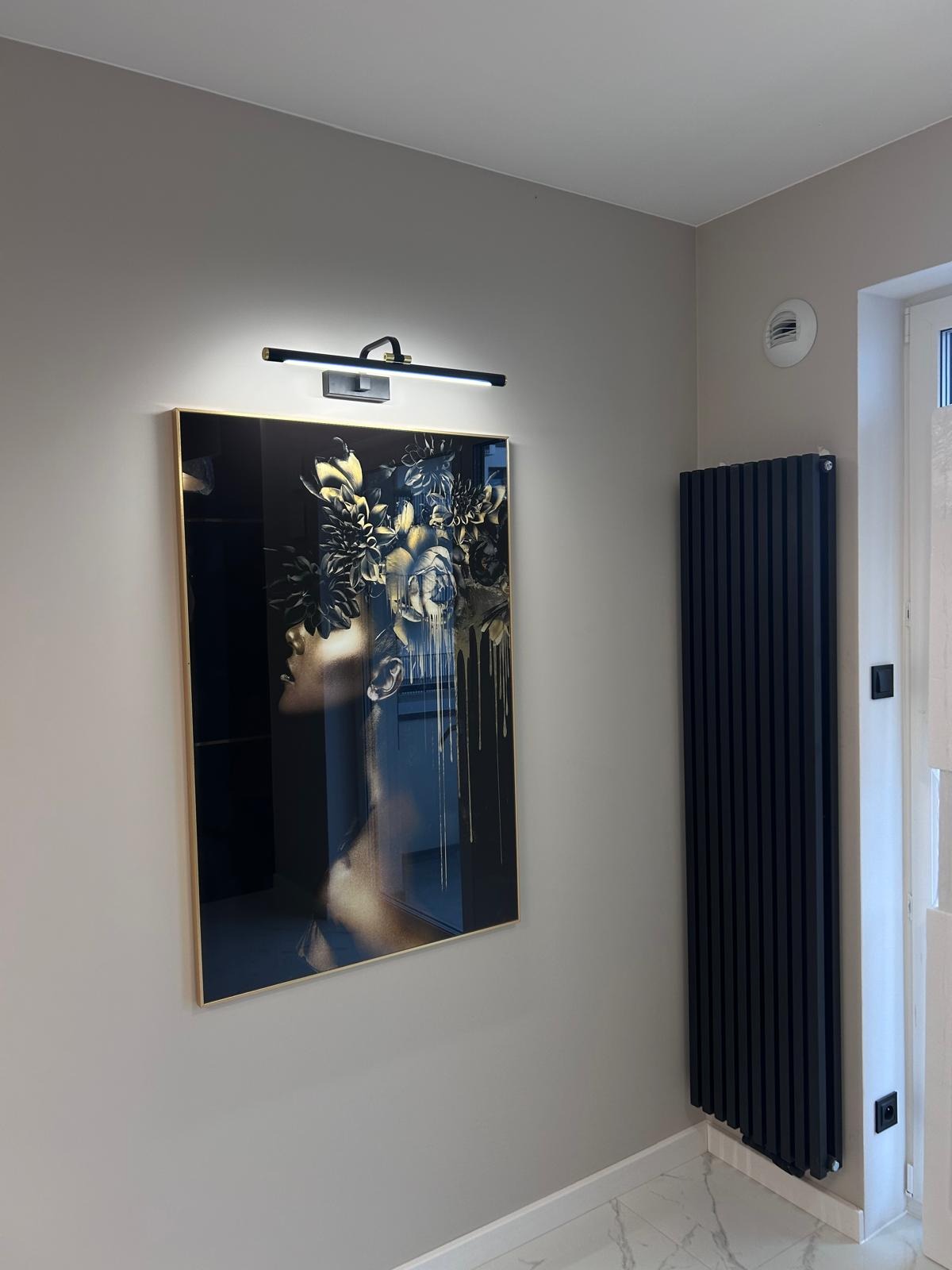 Obraz w złotej ramie z motywem kobiety w kwiatach, oświetlony lampą ścienną, zawieszony na szarej ścianie w korytarzu, obok czarny grzejnik żeberkowy.