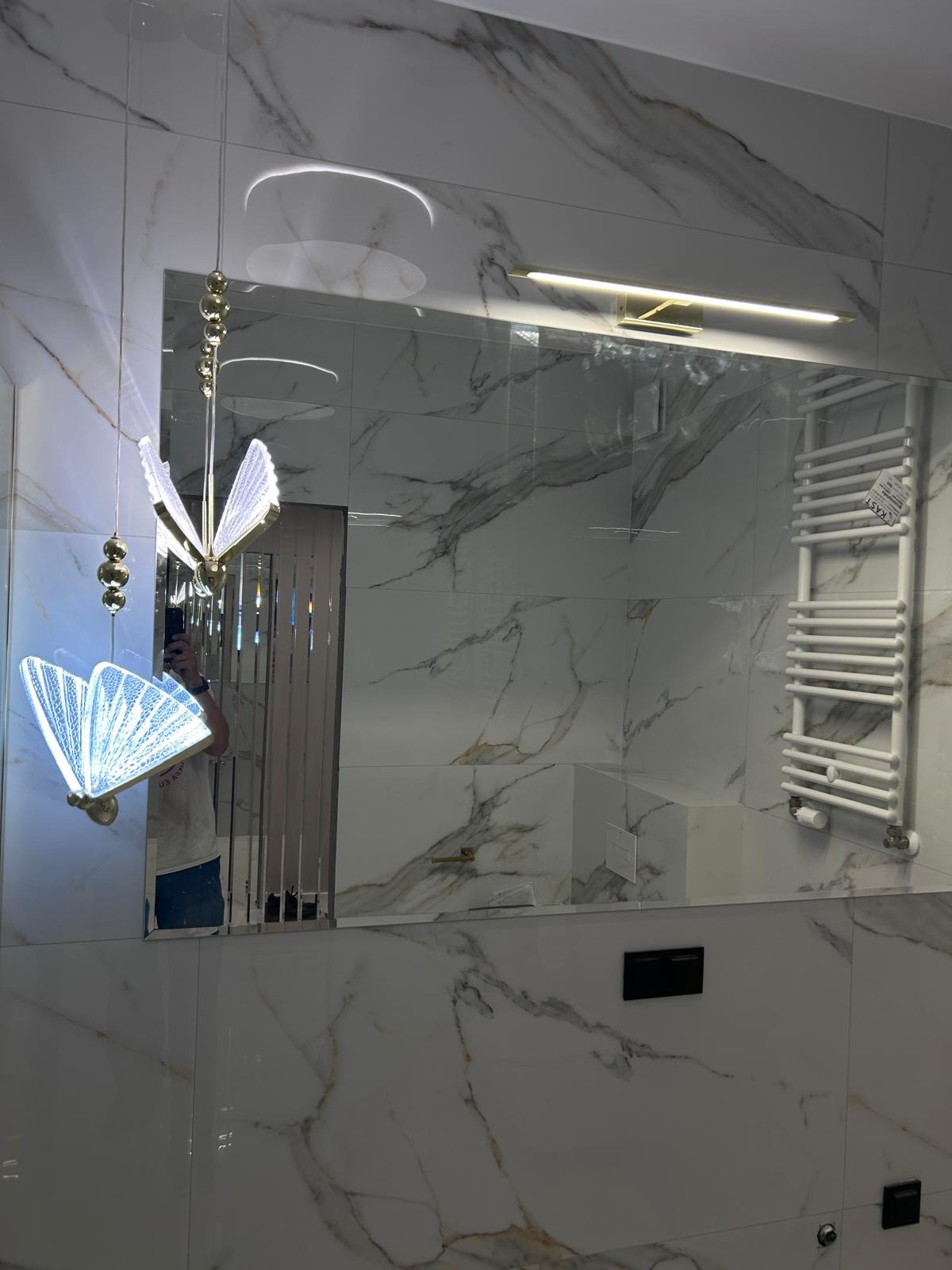 Błyszcząca łazienka z marmurowymi płytkami, dekoracyjnym oświetleniem w kształcie motyli i grzejnikiem drabinkowym, odbicie osoby robiącej zdjęcie w lustrze.