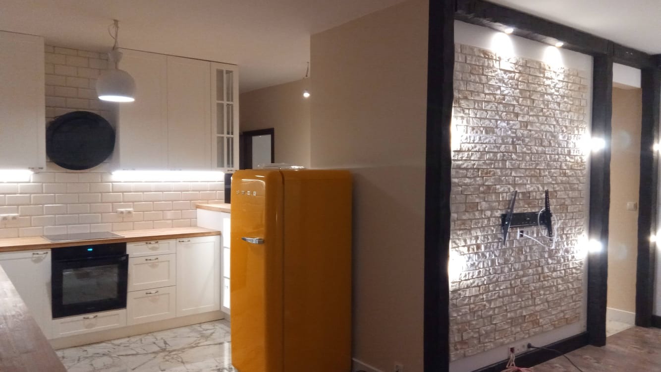 Żółta lodówka Smeg w nowocześnie wykończonej kuchni z białymi szafkami i ceglaną ścianą z zamontowanym uchwytem na telewizor.