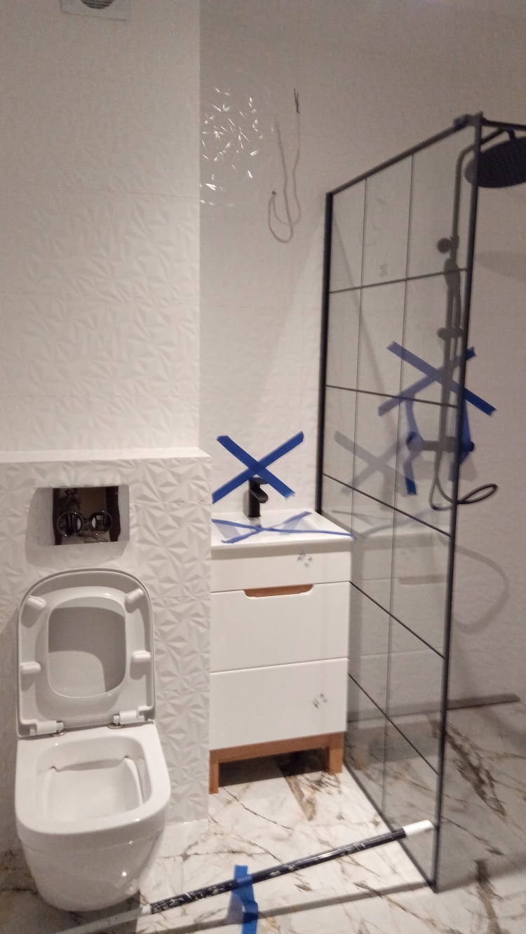 Nowoczesna łazienka w trakcie wykańczania: białe płytki 3D, toaleta podwieszana, szafka z umywalką i czarną armaturą zabezpieczone niebieską taśmą malarską, szklana kabina prysznicowa z czarnymi...