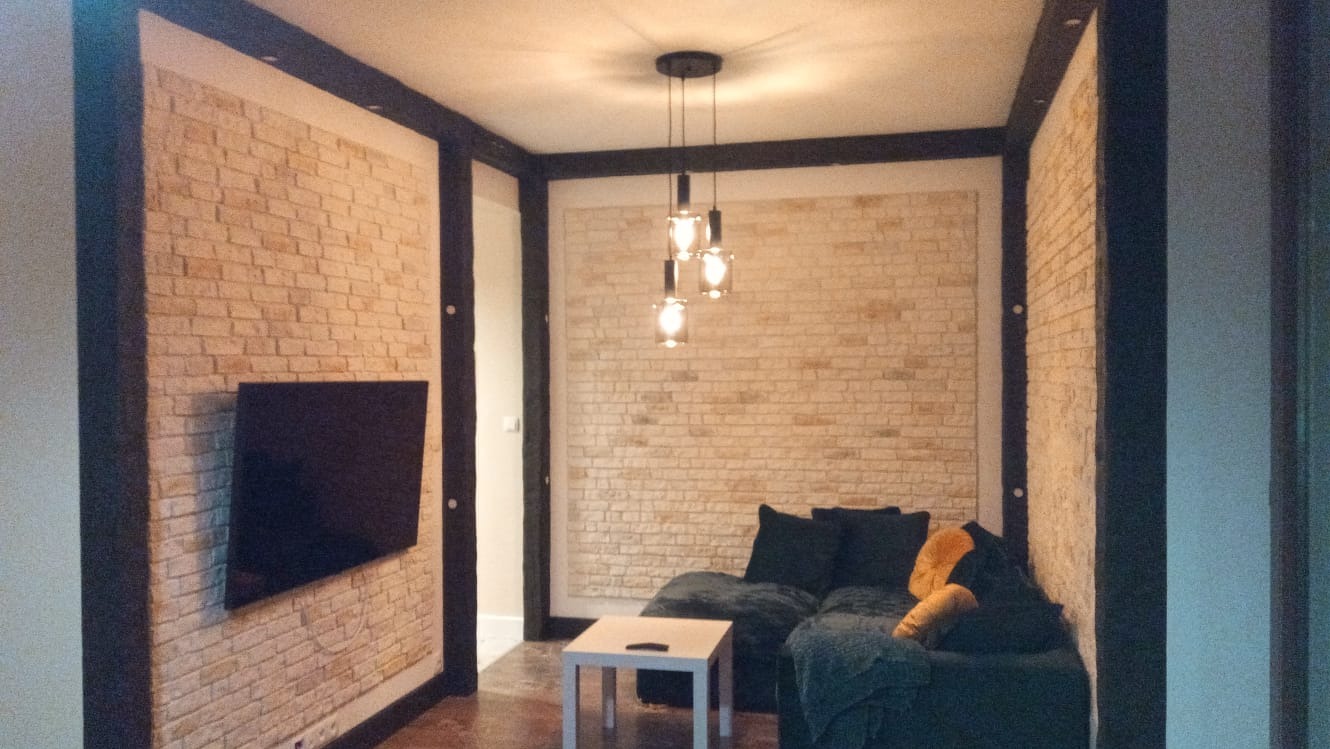 Salon z dekoracyjną ścianą z cegły, obramowaną ciemnym drewnem, na której wisi telewizor; nad sofą wisi nowoczesna lampa z trzema szklanymi kloszami.