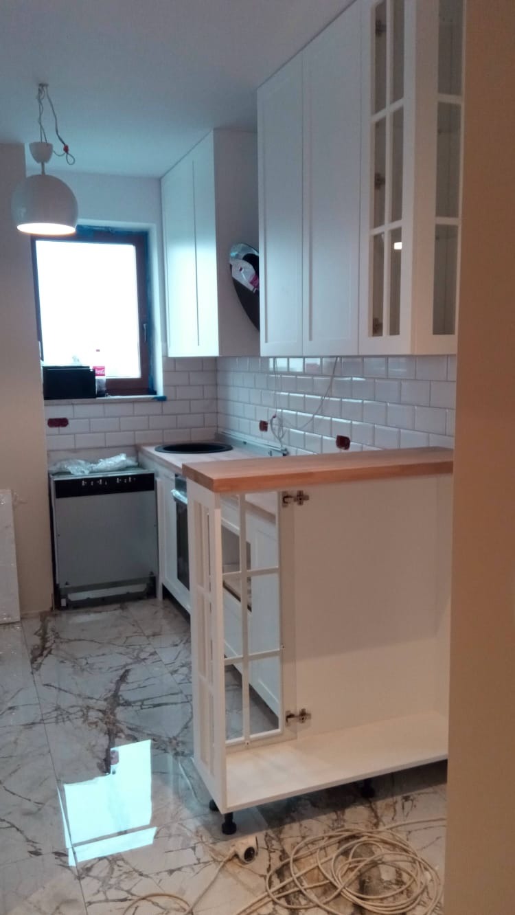 Nowoczesna kuchnia w trakcie wykończenia: białe szafki z drewnianym blatem, marmurowa podłoga, widoczne kable i drobne niedoskonałości.
