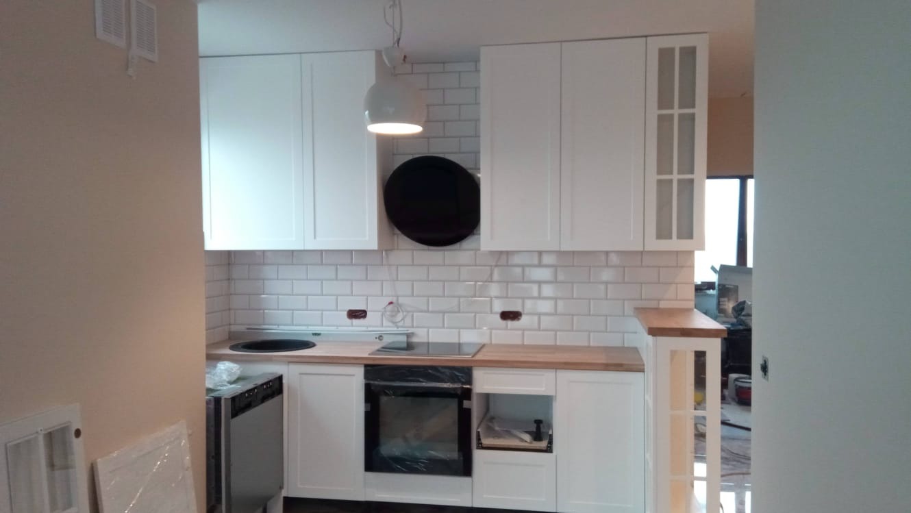 Kuchnia w trakcie wykańczania: białe szafki wiszące i stojące, białe płytki na ścianie, drewniany blat, brakujące listwy przypodłogowe, widoczne przewody elektryczne.