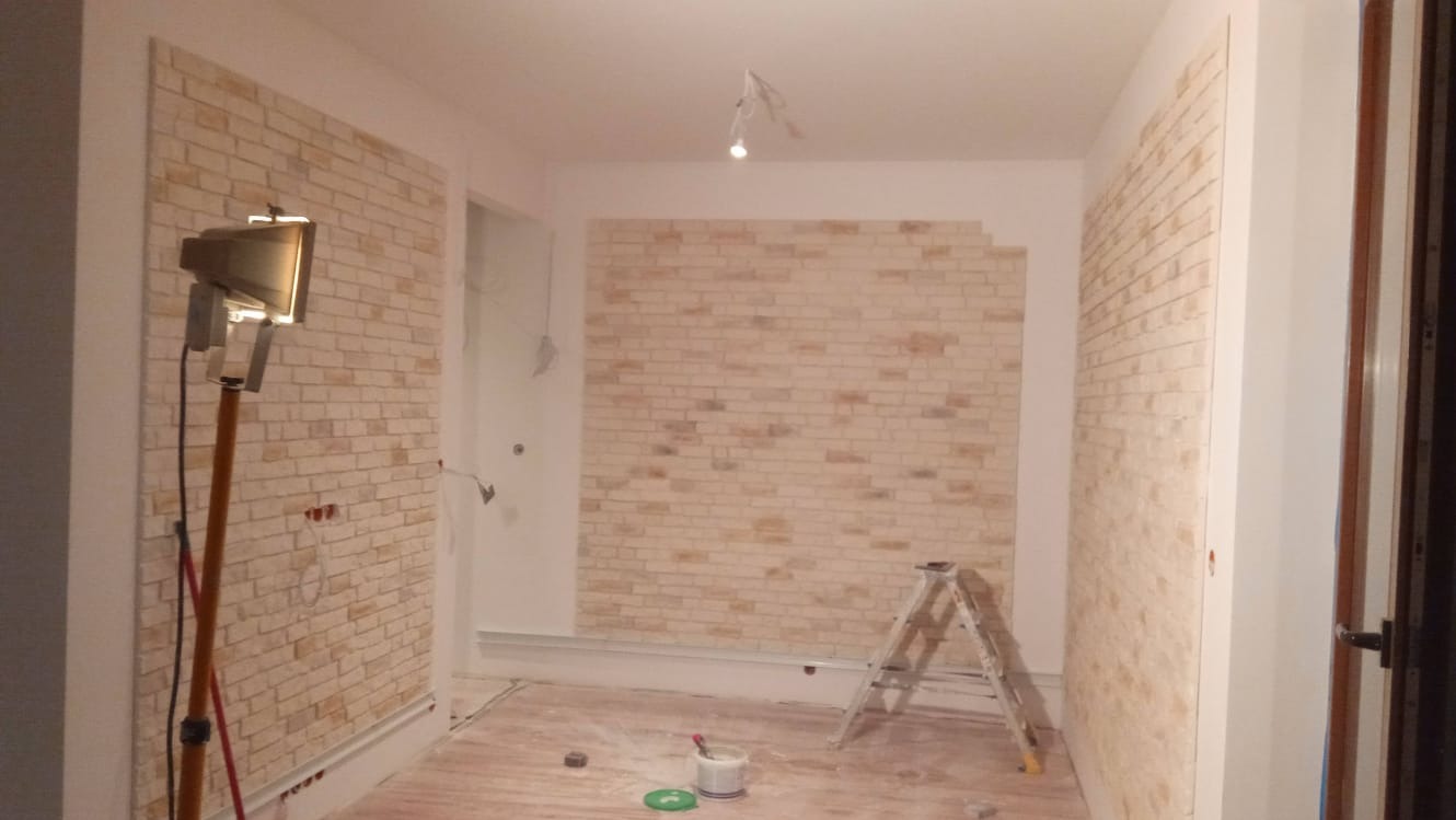 Wnętrze w trakcie prac wykończeniowych: ściany pokryte panelami imitującymi cegłę, ustawiona drabina, widoczne narzędzia i oświetlenie robocze.