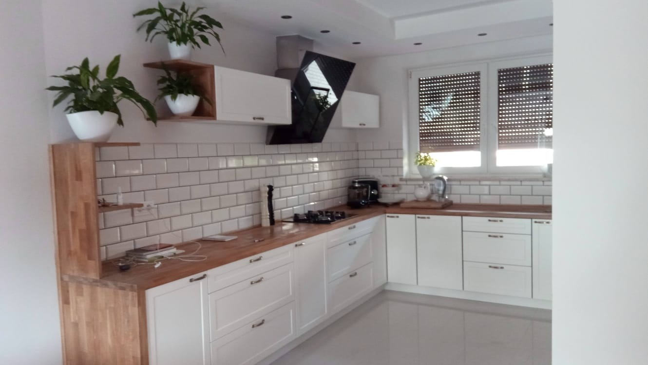 Jasna kuchnia z drewnianym blatem, białymi szafkami i płytkami typu metro, uzupełniona o rośliny doniczkowe i nowoczesny okap.