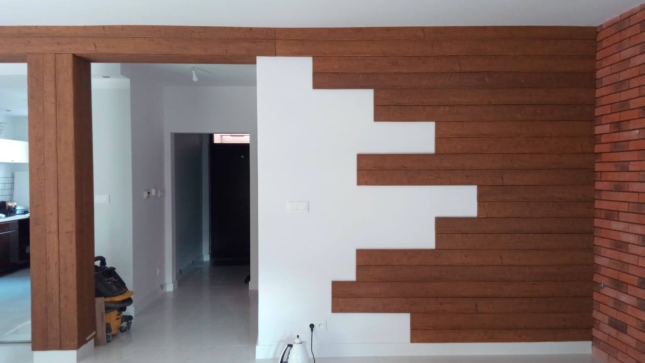 Nowoczesne wykończenie wnętrza z drewnianymi panelami na ścianie, tworzącymi dekoracyjny wzór na białym tle, oraz fragment ściany z cegły w odcieniach czerwieni.