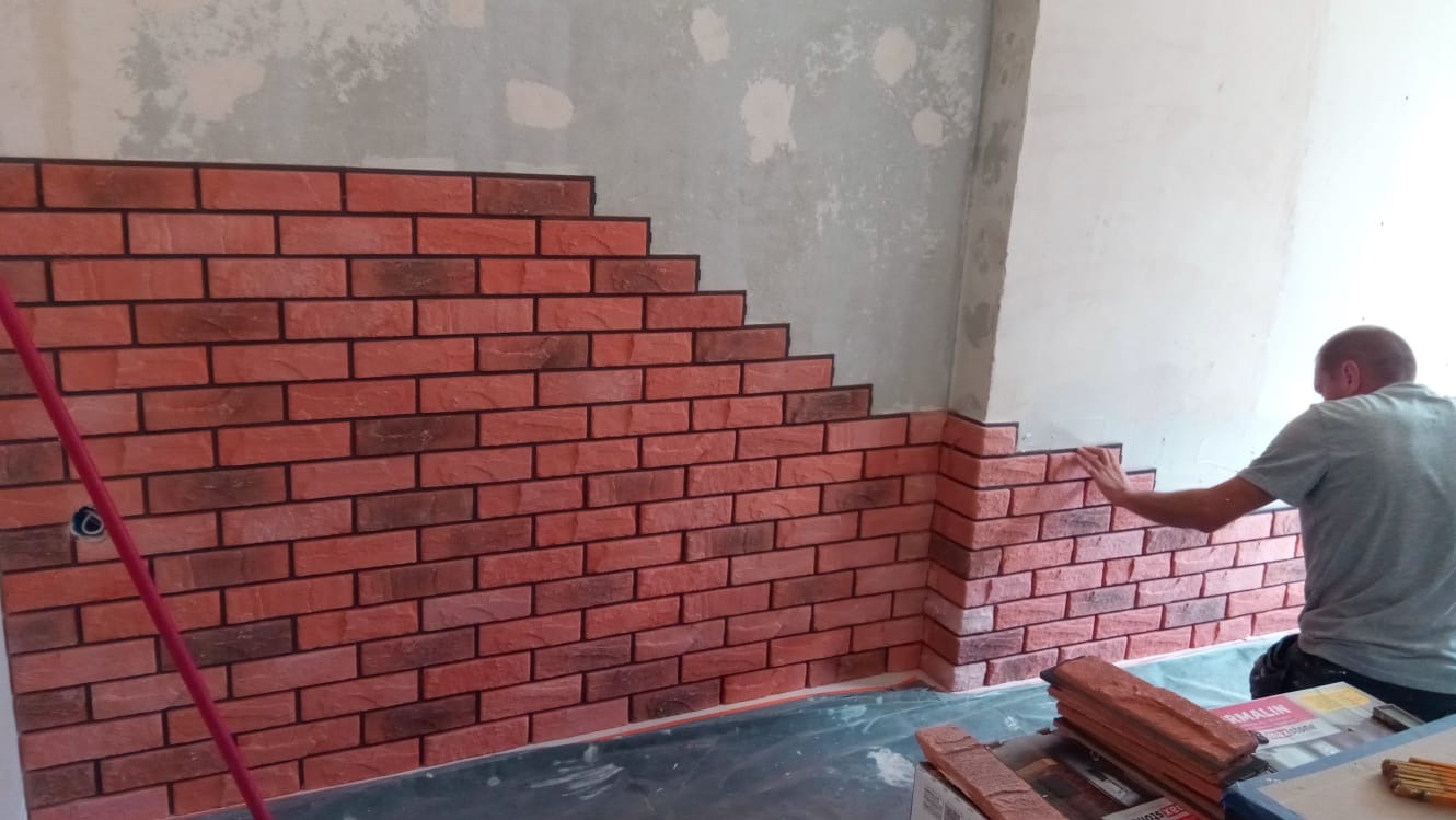 Montaż dekoracyjnej cegły na ścianie wewnętrznej, widoczny fragment ściany surowej i częściowo wykończonej, pracownik dociska element do ściany, widoczne opakowania cegieł i czerwony mop.