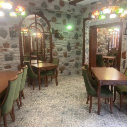 HUGO INVESTMENTS SPÓŁKA Z OGRANICZONĄ ODPOWIEDZIALNOŚCIĄ - Wnętrze restauracji z kamiennymi ścianami, drewnianymi stołami i krzesłami obitymi zielonym aksamitem, oświetlone kolorowymi lampami w stylu tureckim.
