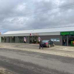 HUGO INVESTMENTS SPÓŁKA Z OGRANICZONĄ ODPOWIEDZIALNOŚCIĄ - Nowoczesny, parterowy budynek handlowo-usługowy z szarą elewacją i białym dachem, widoczne szyldy 'Żabka' i 'Mikro-Cukiernia Głogówek', zaparkowany samochód osobowy i przechodzący mężczyźni.