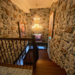 HUGO INVESTMENTS SPÓŁKA Z OGRANICZONĄ ODPOWIEDZIALNOŚCIĄ - Kamienne ściany klatki schodowej z drewnianymi stopniami, oświetlone kryształowym żyrandolem i dekorowane obrazami, widok z góry.