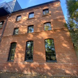 HUGO INVESTMENTS SPÓŁKA Z OGRANICZONĄ ODPOWIEDZIALNOŚCIĄ - Trzykondygnacyjny budynek z czerwonej cegły z czarnymi, prostokątnymi oknami i drzwiami, widziany pod kątem, z opadłymi liśćmi na ziemi.