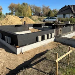 Fundamenty domu w trakcie budowy, z czarną izolacją przeciwwilgociową, widoczne zbrojenia i przygotowany teren, w tle kopiec ziemi i budynek mieszkalny.