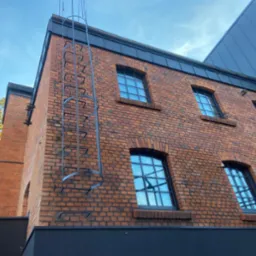 Ceglana elewacja budynku z oknami w czarnych ramach i metalową drabiną przymocowaną do ściany, widok z dołu