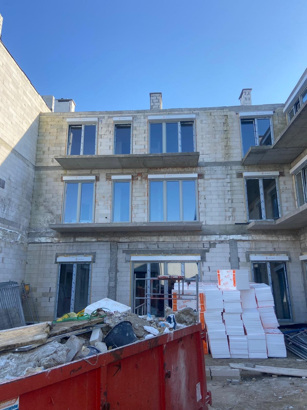 Surowy, trzypiętrowy budynek z bloczków, z zamontowanymi oknami i widocznymi elementami konstrukcyjnymi, w tle błękitne niebo, a na pierwszym planie kontener z odpadami budowlanymi i stos białego...