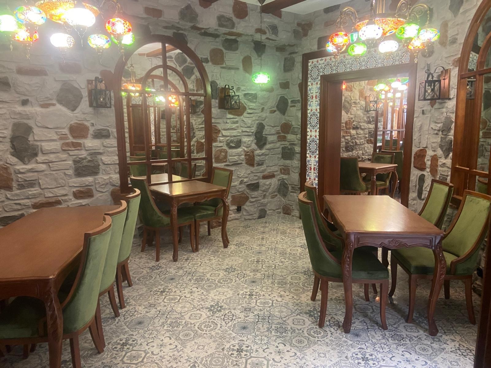 Wnętrze restauracji z kamiennymi ścianami, drewnianymi stołami i krzesłami obitymi zielonym aksamitem, oświetlone kolorowymi lampami w stylu tureckim.